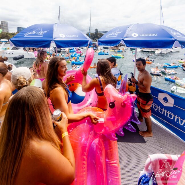 People in swimsuits enjoying a yacht party. A woman carries a pink inflatable unicorn, while others hold drinks. The scene overlooks a harbor with numerous floaties and boats. Several blue umbrellas display the "Liquid IV" logo.