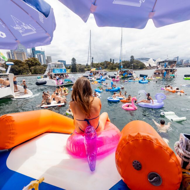 A vibrant scene on a river or lake, with people on colorful inflatable floats enjoying the water. A woman sits on an orange and pink float in the foreground. Other boats and city buildings are visible in the background.