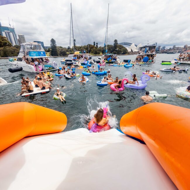 People enjoying a festive gathering on a body of water, with various inflatable rafts and toys. The view is from a large inflatable slide. Boats and a city skyline are seen in the background under a cloudy sky.