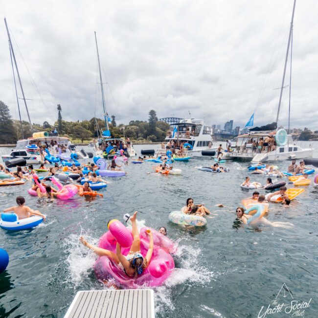 A vibrant scene of people enjoying a boat party, lounging on colorful inflatables in the water. Multiple boats are anchored in the background with people socializing on deck. The sky is cloudy yet bright, setting a festive atmosphere.