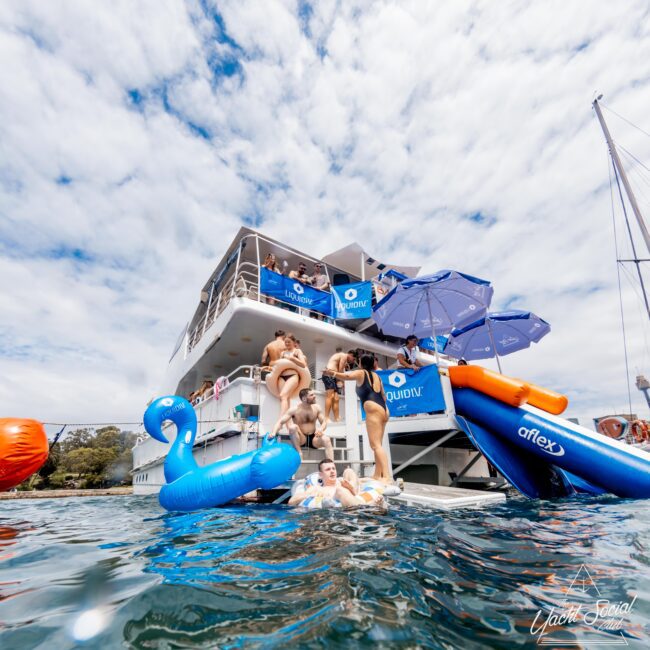 A group of people enjoys a sunny day on a yacht, with some on deck and others in the water. They are using inflatables including a blue swan. The yacht is adorned with blue banners and has a slide extending into the water.