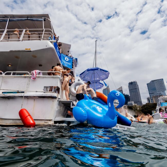 People are enjoying a sunny day on a yacht. Some are swimming or sitting on a large blue inflatable pool float shaped like a swan. The sky is partly cloudy, and buildings are visible in the background.