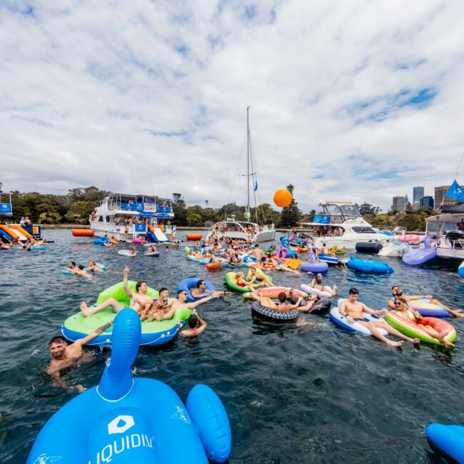 A lively group of people float on inflatable rafts in a bay, surrounded by boats. Various colorful inflatables, including a unicorn and geometric shapes, add vibrancy. The sky is partly cloudy, and city buildings are visible in the background.