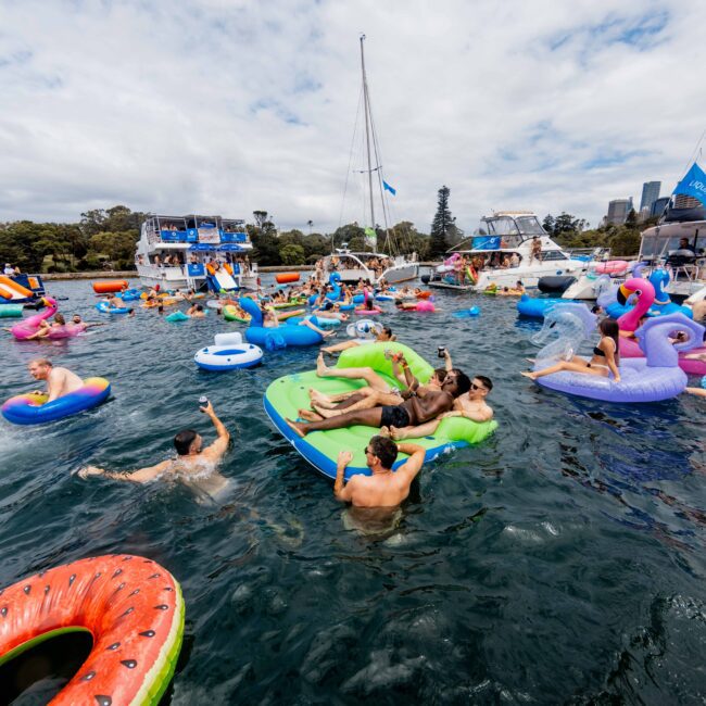 A group of people enjoying a sunny day on colorful inflatable rafts in a body of water. Boats are visible in the background, and the sky is partly cloudy. A watermelon-shaped float is in the foreground.