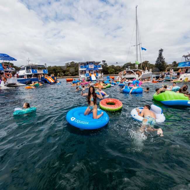 A lively scene of people floating on inflatable rafts in a lake surrounded by boats. The sky is partly cloudy, and participants are enjoying the water on various floaties of different colors and shapes.