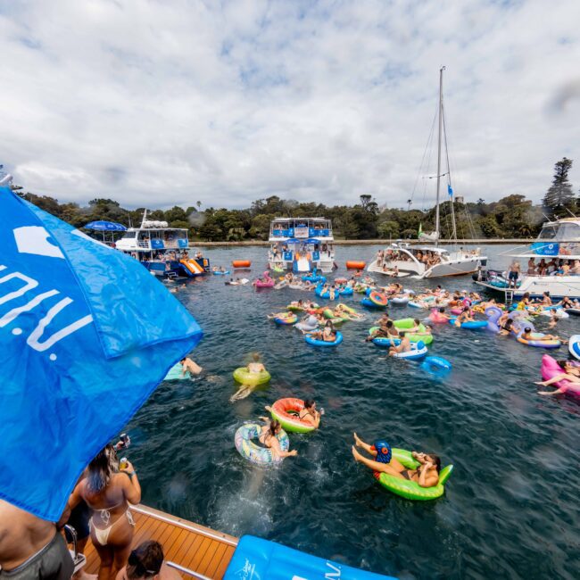 A vibrant scene shows people on colorful inflatable floats having fun in the water. Several boats are anchored nearby with banners and flags. The sky is partly cloudy, and trees are visible in the background.