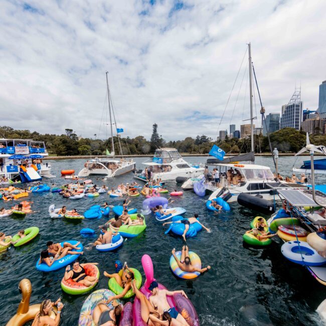 A lively gathering of people on colorful inflatables and boats in a body of water. The scene is festive, with a mix of catamarans and yachts nearby, set against a backdrop of city skyscrapers and a partly cloudy sky.