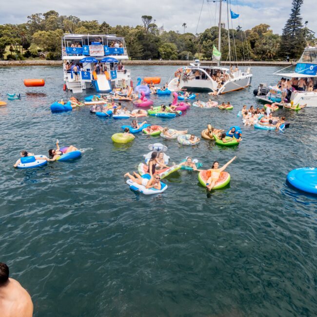 People relaxing on colorful inflatable floats in a lake near several boats. A large yacht with a slide is visible in the background, surrounded by trees and under a partly cloudy sky.