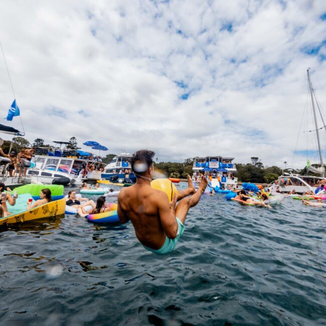 A person mid-flip above a body of water surrounded by others on inflatable floats. Several boats and people are visible in the background under a partly cloudy sky. A "Yacht Social Club" logo is in the corner.