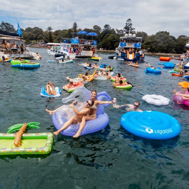 People relaxing on colorful inflatable floats, including a unicorn and a pineapple, in a lake. Boats are anchored nearby, and trees line the shore under a cloudy sky. The mood is festive and carefree.