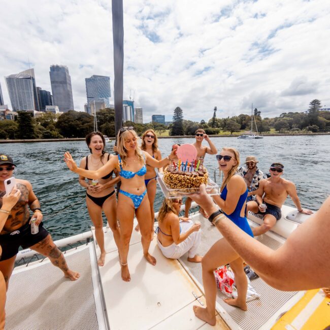 A group of people on a yacht celebrate with a birthday cake. They are smiling and wearing swimsuits. The city skyline is visible in the background under a partly cloudy sky.