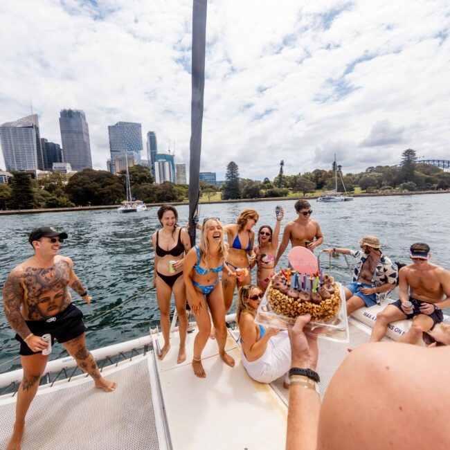 A group of people on a boat celebrating, with some in swimwear. One person is holding a cake with candles. The backdrop features city buildings and a green landscape, suggesting a lively, sunny day by the water.