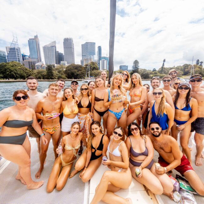 A group of people in swimwear smiling and posing on a boat. The city skyline and trees are visible in the background. It appears to be a sunny day, and the mood is festive and cheerful.