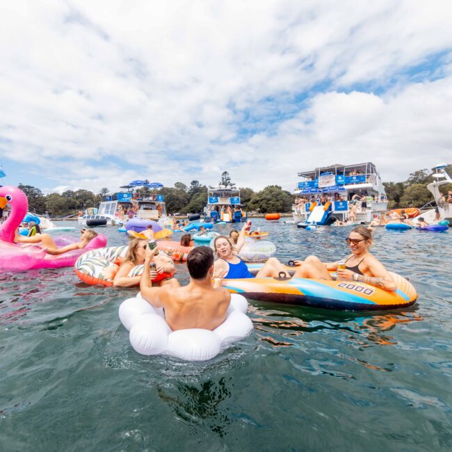 A group of people are relaxing on inflatable floats, including a flamingo and a unicorn, in a body of water. Boats are anchored nearby, and the sky is partly cloudy. The scene is lively and festive.