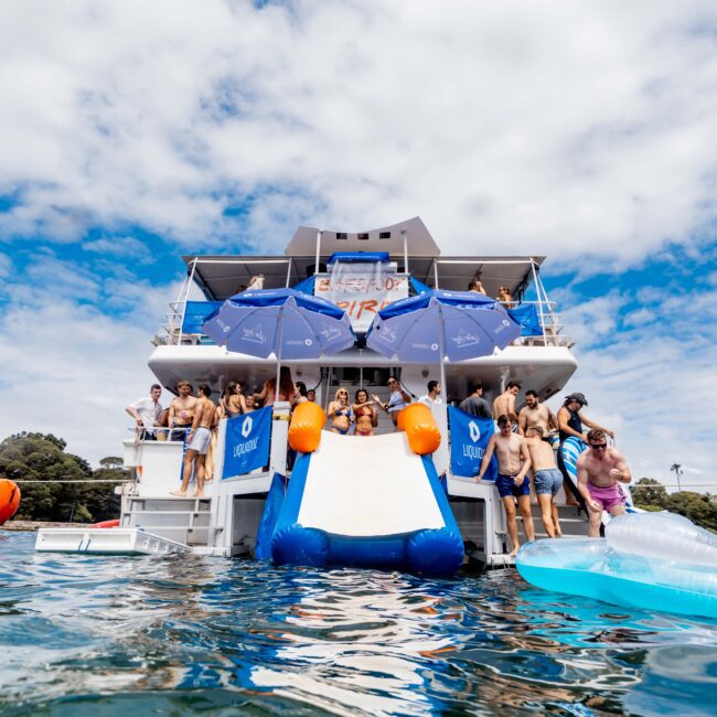 A group of people enjoying a boat party with a slide leading into the water. The boat is adorned with blue umbrellas and there's a festive atmosphere. The sky is partly cloudy, and the water is calm.