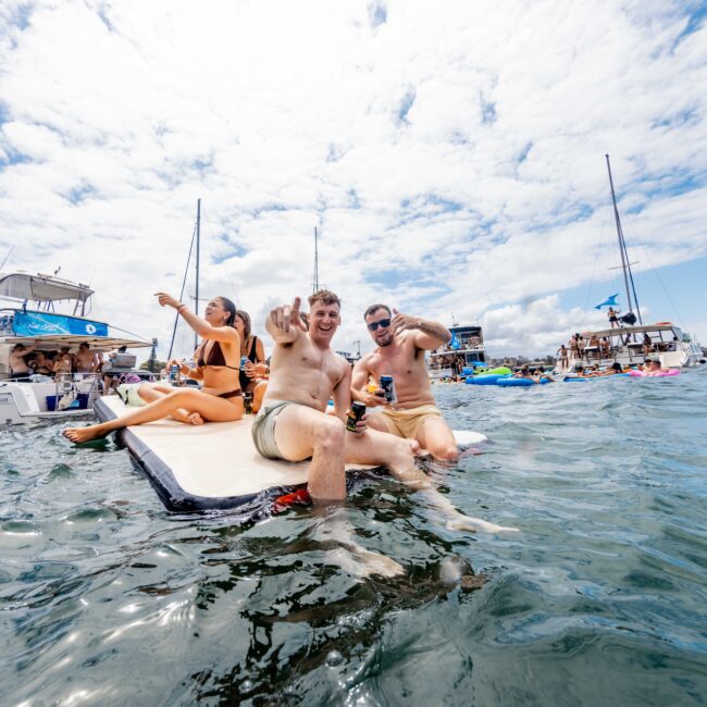 Three people in swimwear sit on a floating mat in the water, smiling and making peace signs. Several boats and inflatable items are in the background under a partly cloudy sky.