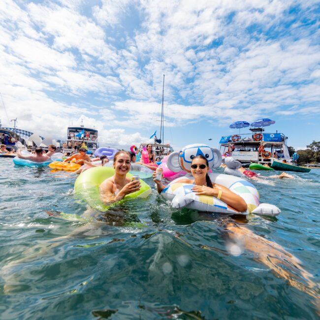 Two people float on inflatable rings in the water, smiling and holding drinks. They are surrounded by others enjoying a sunny day on the water, with boats and a partly cloudy sky in the background.