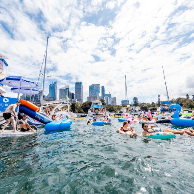 People enjoying a lively floating party on a city waterfront, surrounded by colorful inflatables and boats. Skyscrapers are visible in the background under a partly cloudy sky.