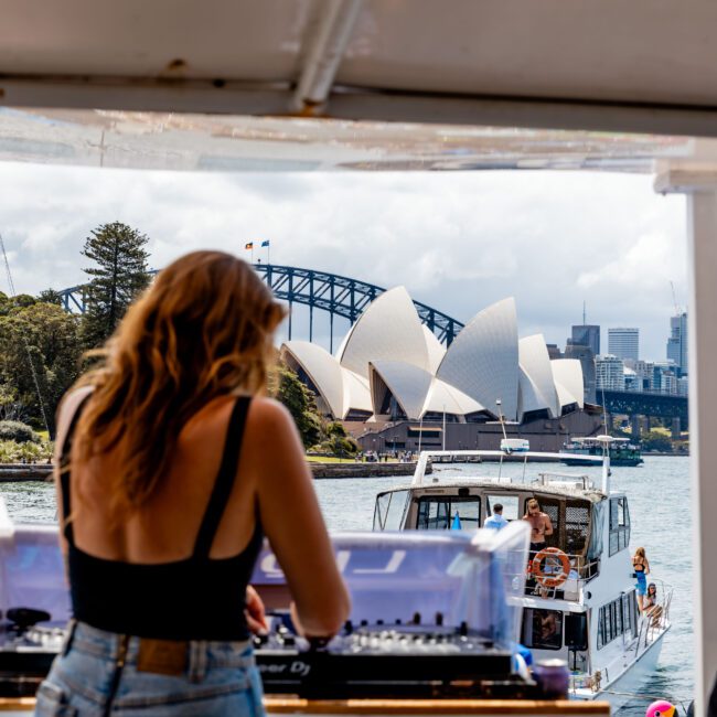 Woman with long hair, wearing a black tank top and denim shorts, DJs on a boat. The Sydney Opera House and Harbour Bridge are visible in the background across the water. A ferry passes by under a partly cloudy sky.