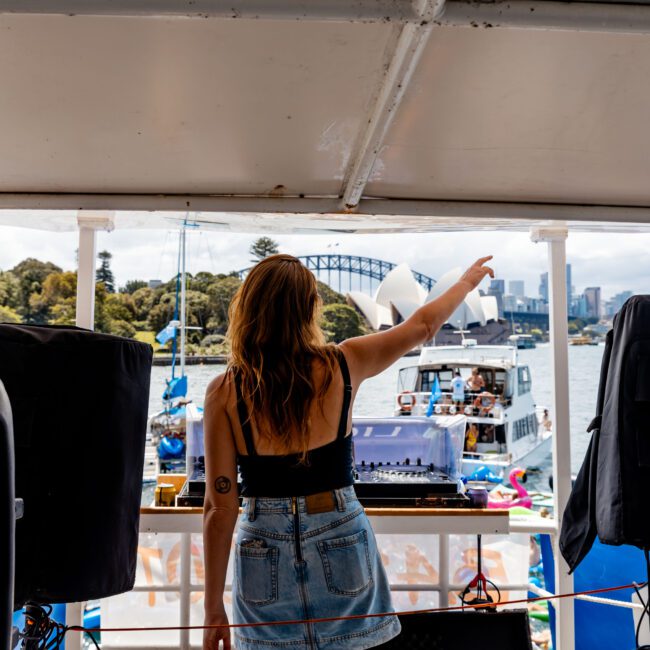 A woman stands on a boat deck, facing away, pointing towards other boats on the water. She's wearing a black tank top and denim shorts. The background features a scenic view of trees and a distant arch bridge under a cloudy sky.