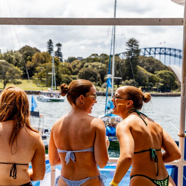 A group of women in swimwear stand on a boat, smiling and talking. In the background, there are boats on the water and a view of the Sydney Opera House and Sydney Harbour Bridge. The scene is sunny and lively.