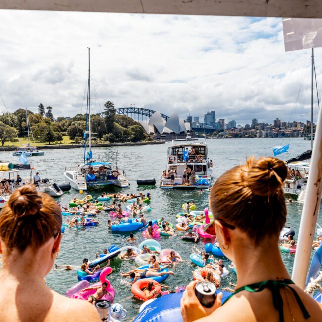 A lively scene with numerous people on colorful floaties in a body of water, enjoying a festive gathering. A boat is nearby with people on board. In the background, the Sydney Opera House and Harbour Bridge are visible under a partly cloudy sky.