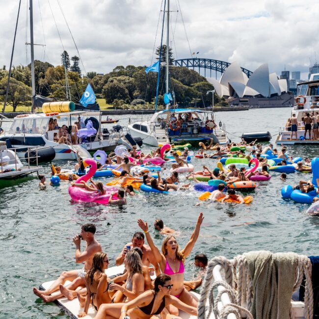 A lively scene on a sunny day with people enjoying a boat party. Many are on colorful inflatable floats in the water. Boats are docked nearby, with a bridge and the Sydney Opera House visible in the background.