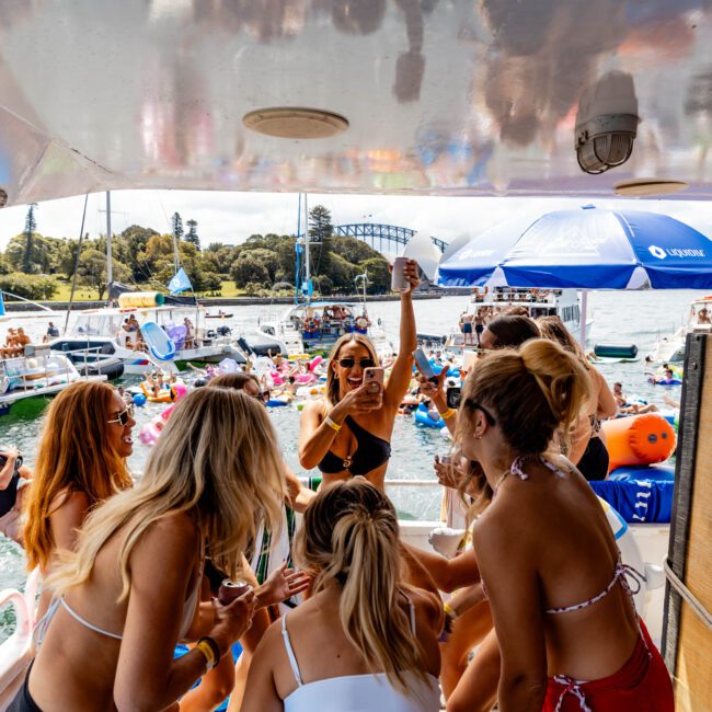 A group of people in swimwear enjoy a party on a boat, surrounded by other boats and colorful inflatables on the water. A bridge and trees are visible in the background. Some people are taking photos and interacting energetically.