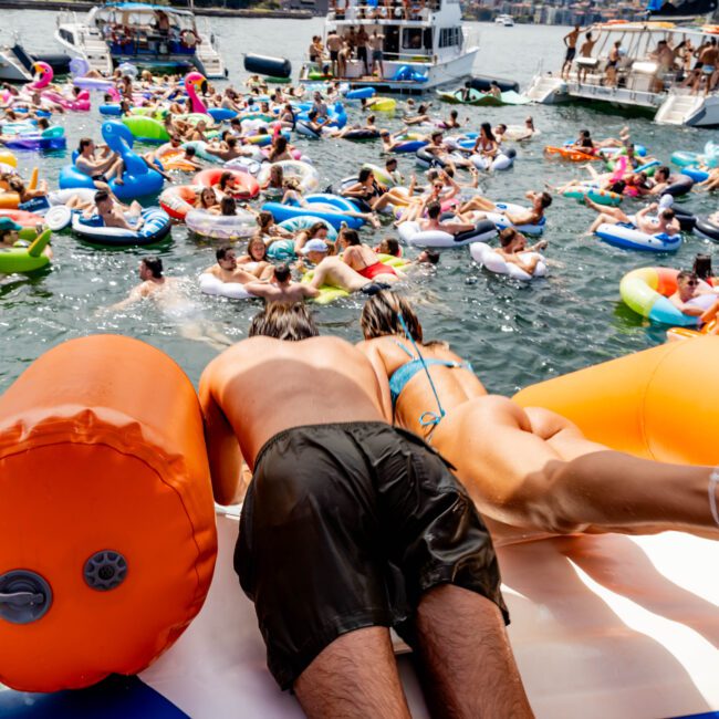 A crowded lake scene with numerous people on colorful inflatable tubes and floats. Two individuals in swimwear are in the foreground on a large inflatable structure. Boats with passengers are in the background under a sunny sky.