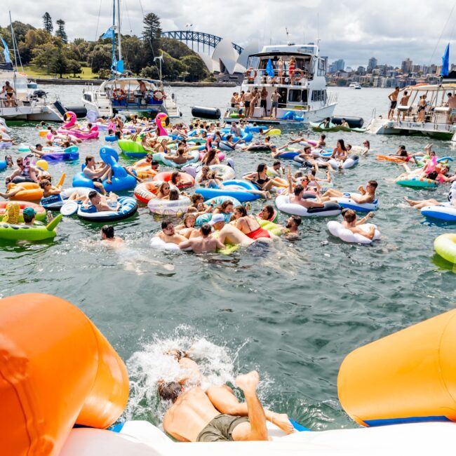 A large group of people relax on colorful inflatable pool floats in a sunny harbor. Boats are anchored nearby, and the skyline, including a bridge, is in the background. The atmosphere is lively with a festive water gathering.