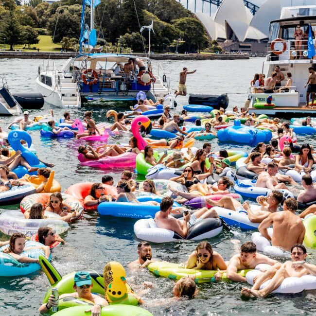 A group of people on colorful inflatable rafts and floats enjoy a sunny day on the water near Sydney Opera House and Harbour Bridge. Boats are anchored nearby, and the scene is lively and festive.