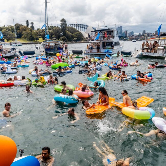 A lively scene of people enjoying a party on a waterfront. Many are in colorful floaties in the water, surrounded by anchored boats. The skyline and a bridge are visible in the background under a partly cloudy sky.