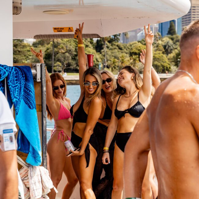 Group of five women in swimwear smiling and posing for a photo on a yacht. They are holding drinks and making playful gestures. The yacht is docked, with water and a cityscape visible in the background.