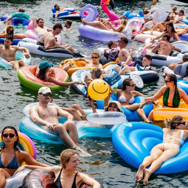 A large group of people relax on colorful inflatable floats in a body of water. Some floats are shaped like animals, including unicorns and ducks. Boats are docked in the background. It's a sunny day, and everyone appears to be enjoying themselves.