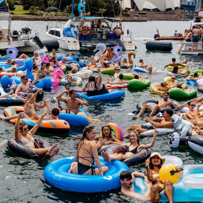 A lively group of people enjoy a sunny day on Sydney Harbour, floating on colorful inflatables. Boats are anchored nearby, with the Sydney Opera House and Harbour Bridge visible in the background. The atmosphere is festive and vibrant.