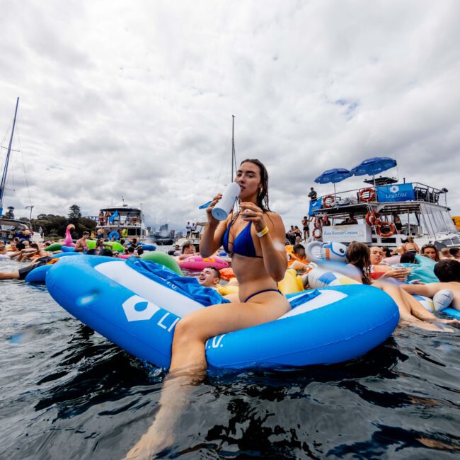 Woman in a blue bikini sits on a blue inflatable float, drinking from a can. She's surrounded by people on various inflatables in a body of water. Boats and a cloudy sky are in the background.