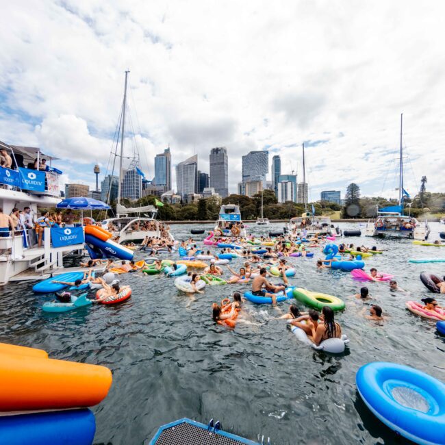 A lively scene of people enjoying a floating party in a city harbor. Numerous individuals are on colorful inflatable rings and floats. A party boat is on the left, and skyscrapers can be seen against a partly cloudy sky in the background.