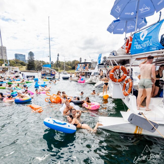 People are enjoying a party on a yacht and in the surrounding water. They are on floaties of various bright colors, with more boats in the background. The yacht has umbrellas and life preservers on board. Buildings and trees are visible in the distance.