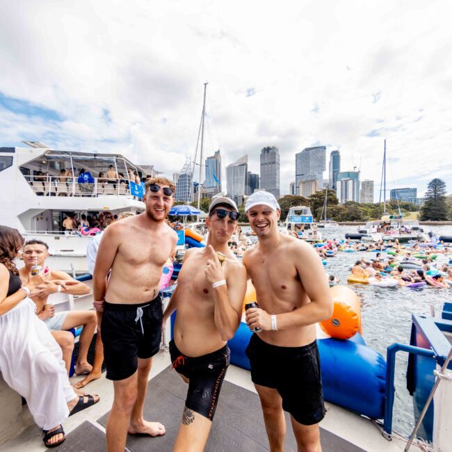 Three men in swim gear pose playfully on a boat. The background shows a crowded water scene with people on inflatables and other boats, set against a city skyline under a partly cloudy sky.