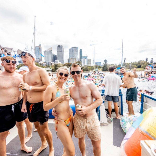 A group of people enjoying a boat party. A woman in a colorful bikini and sunglasses stands with a man in a light shirt and shorts, both smiling and holding drinks. The background shows a city skyline and water inflatables.