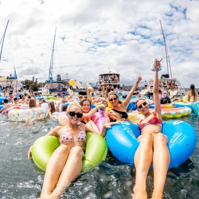 A group of people relax on colorful inflatable rings in the water, surrounded by other swimmers and boats. The sky is partly cloudy. The scene is lively, with many people enjoying the event.