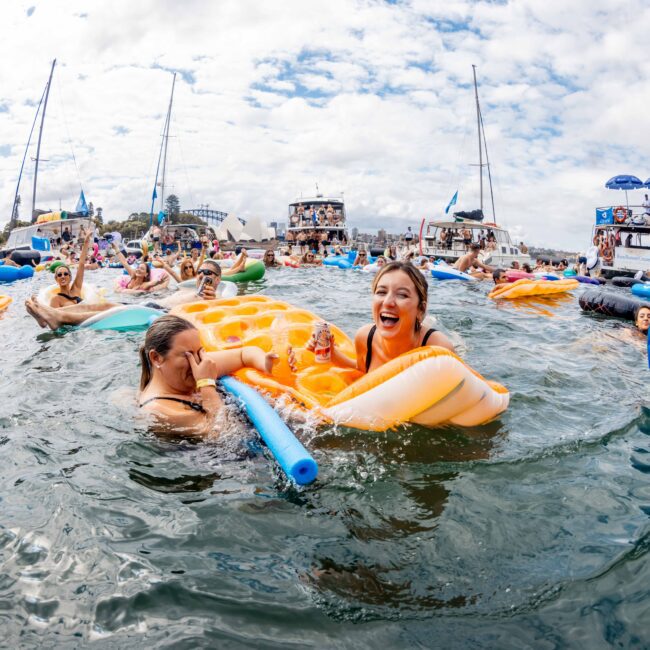 A lively scene of people enjoying a floating pool party on a sunny day. Individuals are on inflatable loungers and floats, chatting and laughing. Boats are docked in the background, and the sky is partly cloudy.