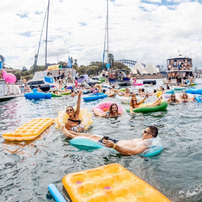 A lively scene of people on inflatable rafts in a harbor, surrounded by boats. Various colorful floats are in the water, with a view of a bridge and skyline in the background. The atmosphere is festive and relaxed.