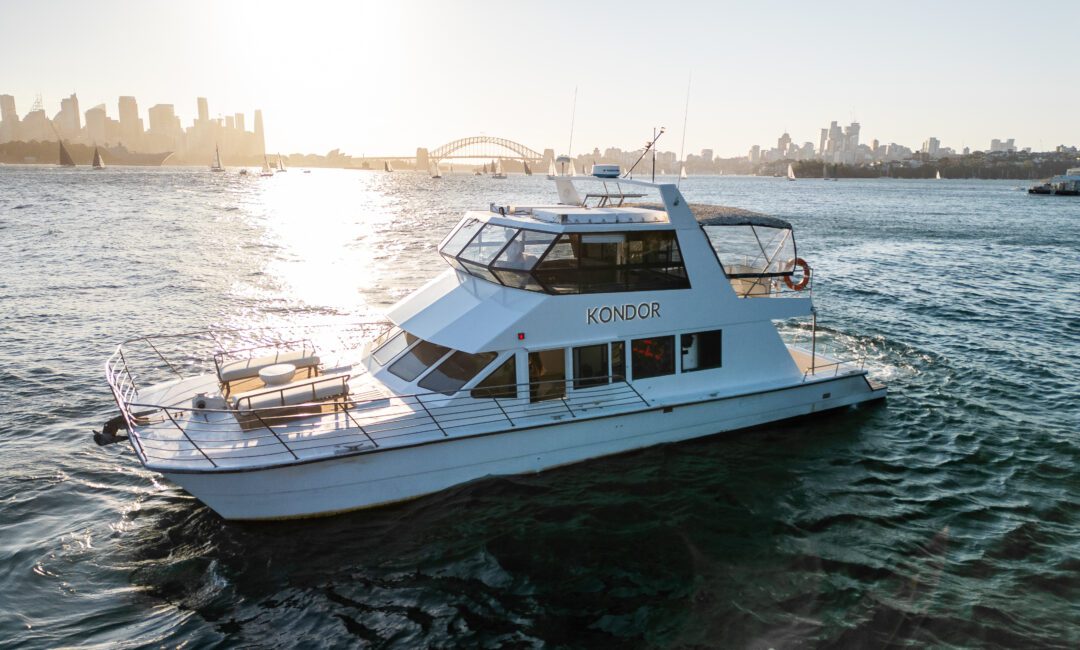 A white boat named "Kondor" on a body of water, cruising toward the camera. The sun sets in the background, casting a warm glow and reflecting on the water. City buildings and a bridge are visible on the horizon.