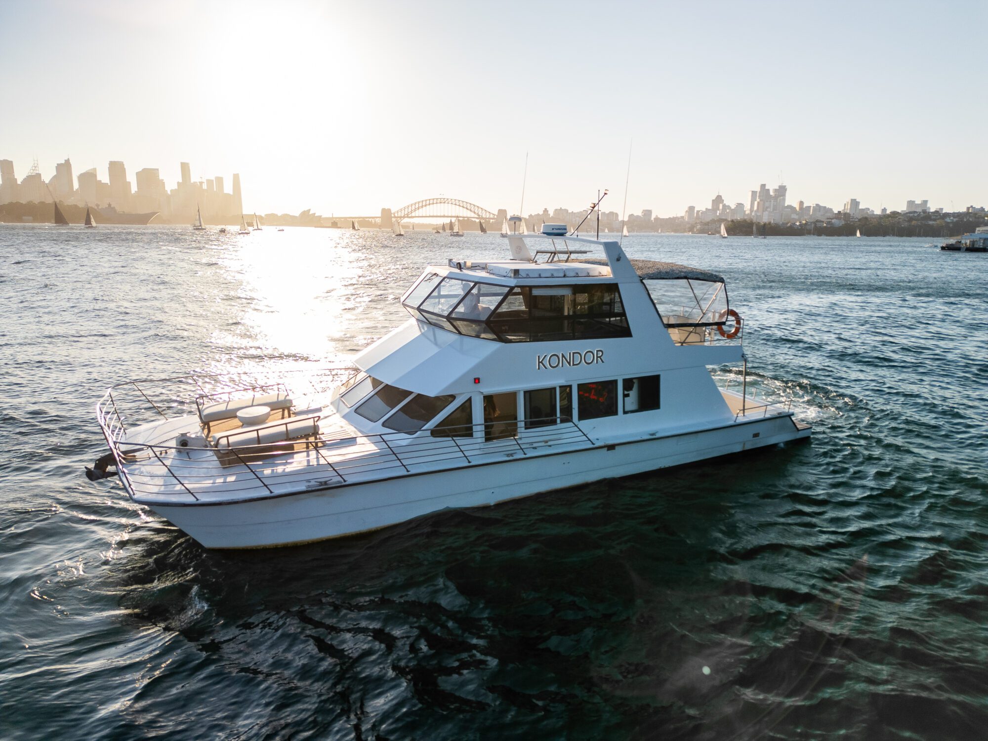 A white boat named "Kondor" on a body of water, cruising toward the camera. The sun sets in the background, casting a warm glow and reflecting on the water. City buildings and a bridge are visible on the horizon.