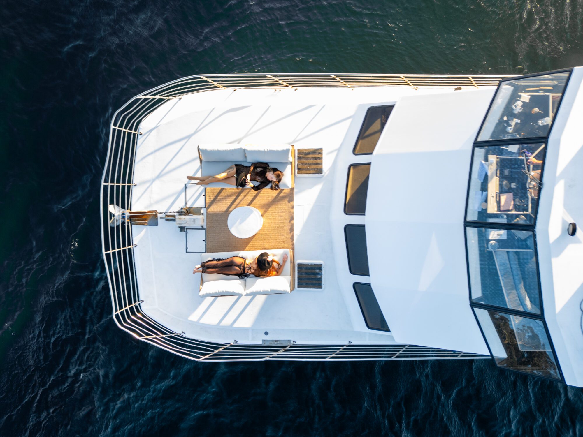 Aerial view of two people relaxing on the deck of a white yacht. They are lying on sun loungers with cushions, positioned around a small round table. The yacht is surrounded by dark blue water, suggesting a calm day at sea.