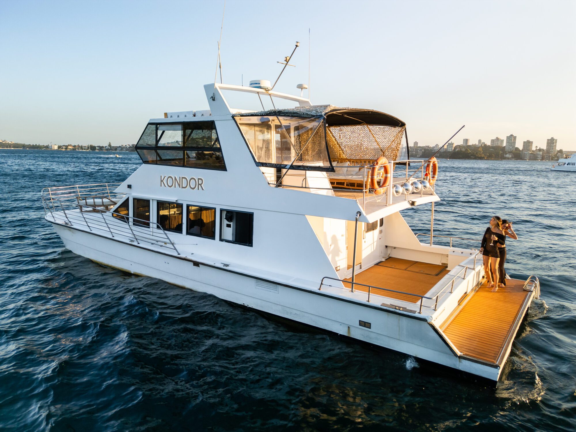 A white yacht named "Kondor" is sailing on the ocean. The vessel has a sleek design with orange deck flooring. Two people stand on the back deck, taking in the view. The skyline of a city is visible in the distance under a clear sky.