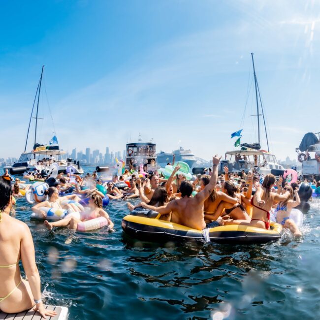A lively scene of people on inflatable rafts and boats in a body of water with a city skyline in the background. The group is enjoying sunny weather, with many individuals socializing and relaxing under clear blue skies.