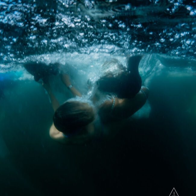 Two people swimming underwater in a dark, wavy sea. Sunlight filters through the water, illuminating their movements. The lower half of the image displays ripples on the surface, while the figures are partially silhouetted beneath.