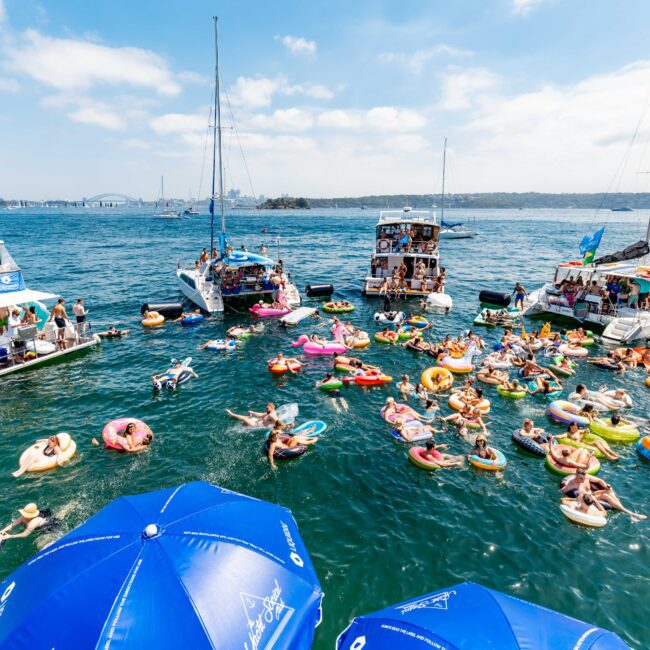 A lively scene of people relaxing on colorful inflatables in the water between several boats on a sunny day. The boats are anchored near each other, and the sky is clear with a few clouds. Some large blue umbrellas are visible in the foreground.